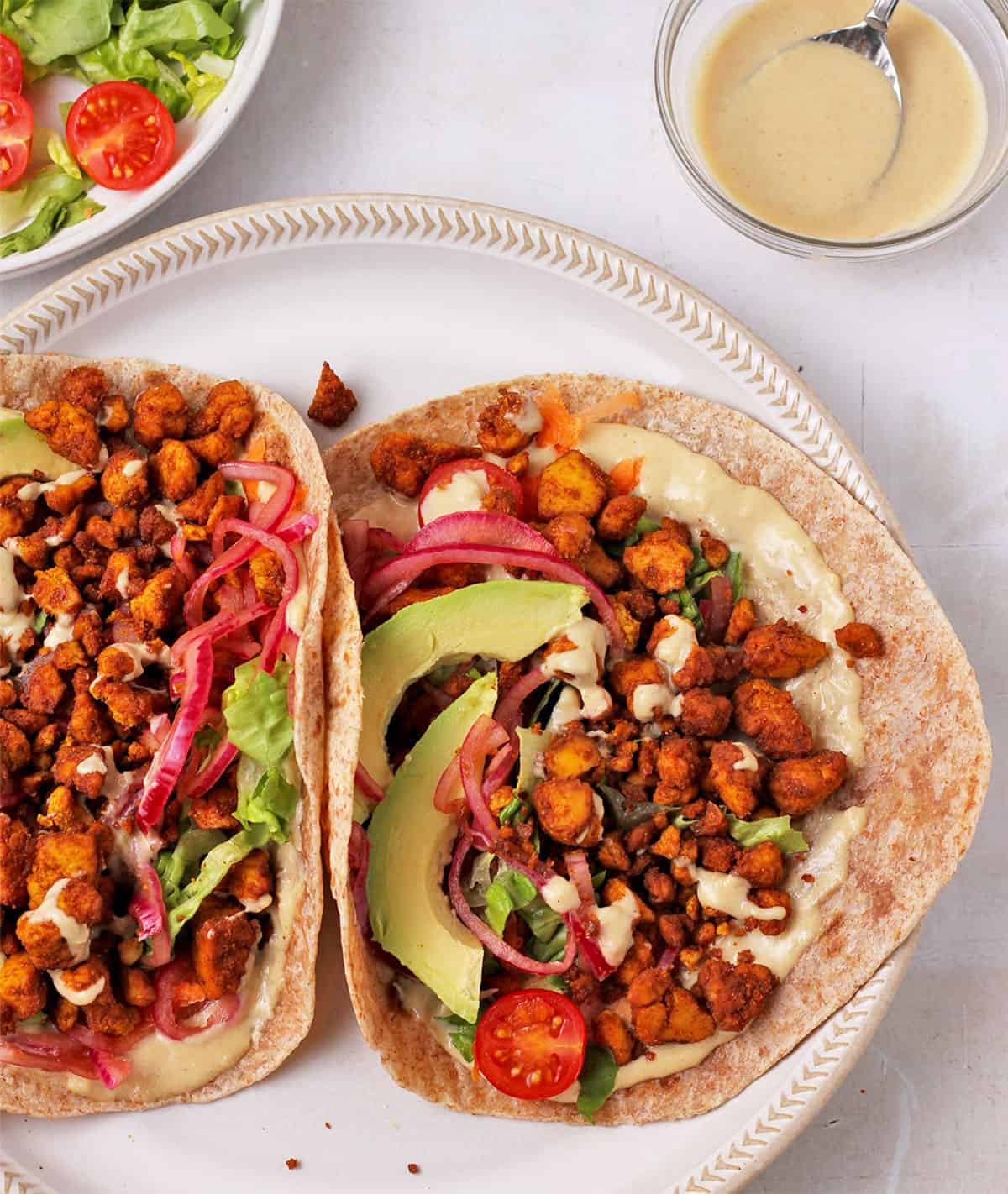 Two wraps with crumbled tofu, tomatoes, avocado, lettuce, pickled red onions, and tahini dressing.