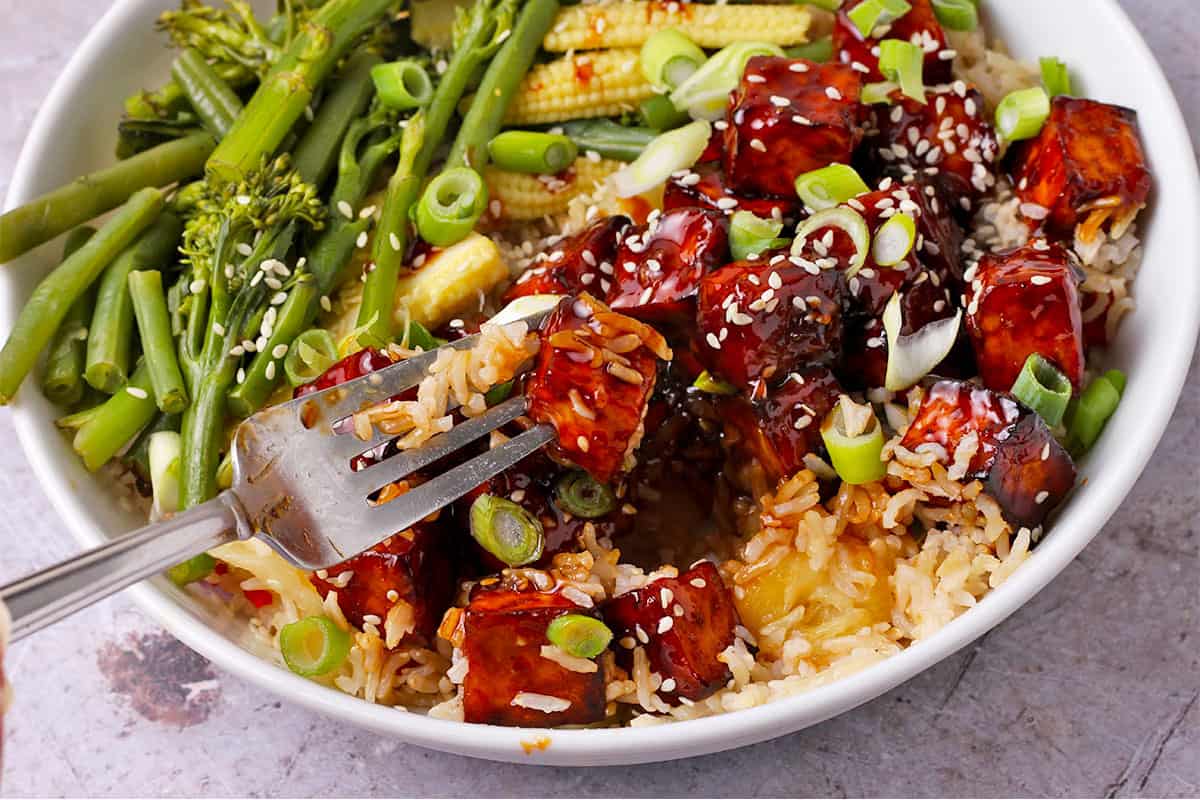 A fork holds a cube of teriyaki tofu over a bowl of tofu with rice, vegetables, and sauce.