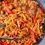 Gochujang udon noodles with cabbage, red peppers, tofu, and sesame seeds and tongs in a pan.