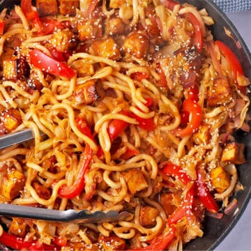 Gochujang udon noodles with cabbage, red peppers, tofu, and sesame seeds and tongs in a pan.