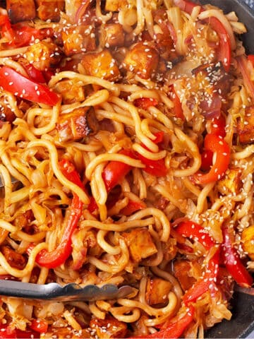 Gochujang udon noodles with cabbage, red peppers, tofu, and sesame seeds and tongs in a pan.