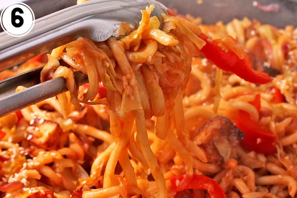 Gochujang noodles are lifted with tong over a pan of noodles, vegetables, and tofu.