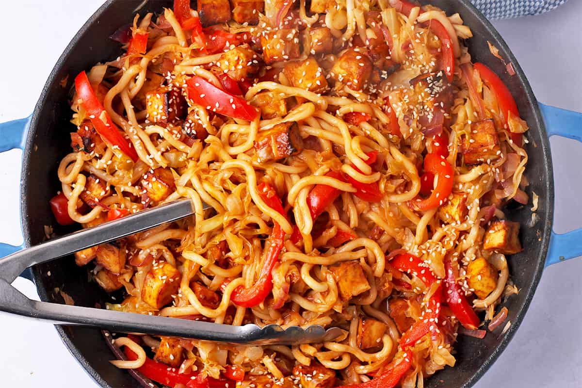 A pan of gochujang noodles with tofu and vegetables with tongs.